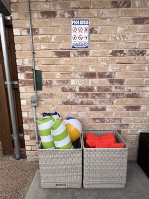 A brick wall with a pool rules sign and two wicker baskets containing beach balls and a red towel.