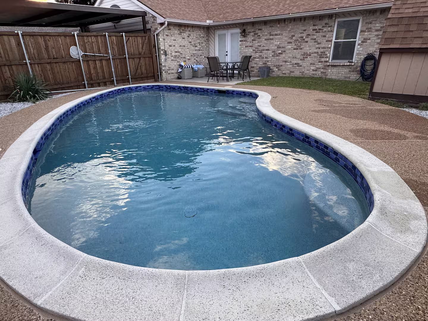 A swimming pool with a curved edge, surrounded by a stone deck and a wooden fence.