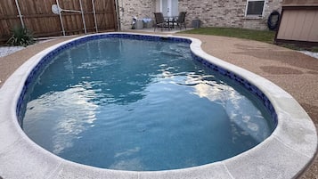 A swimming pool with a curved edge, surrounded by a stone deck and a wooden fence.