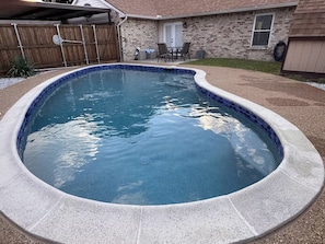 A swimming pool with a curved edge, surrounded by a stone deck and a wooden fence.