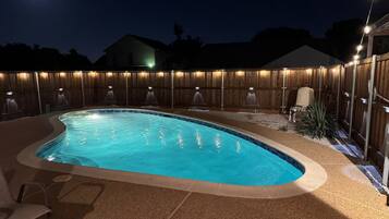 A swimming pool with a wooden fence and lights, surrounded by a patio area.