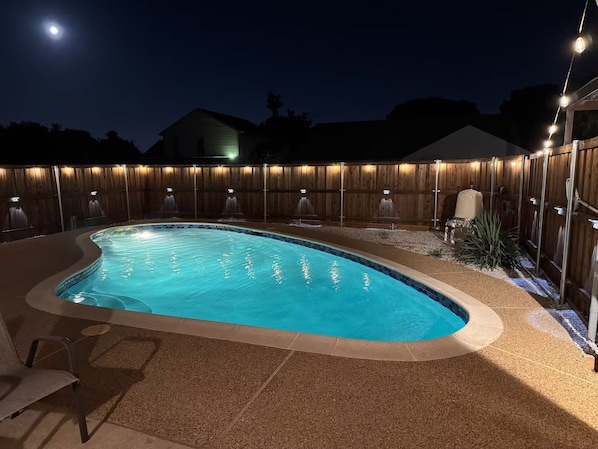 A swimming pool with a wooden fence and lights, surrounded by a patio area.