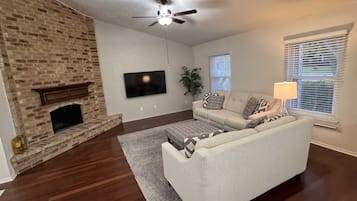 A living room with a brick fireplace, a ceiling fan, a flat-screen TV, a sofa, and a window with blinds.
