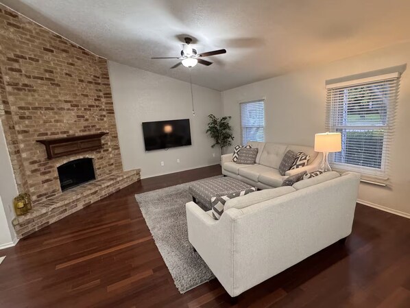 A living room with a brick fireplace, a ceiling fan, a flat-screen TV, a sofa, and a window with blinds.