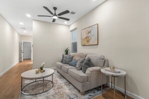 A living room with a ceiling fan, a sofa with cushions, a coffee table, and a side table with a decorative item. - Modern 3BR Houston Retreat Stylish & Spacious (Houston)