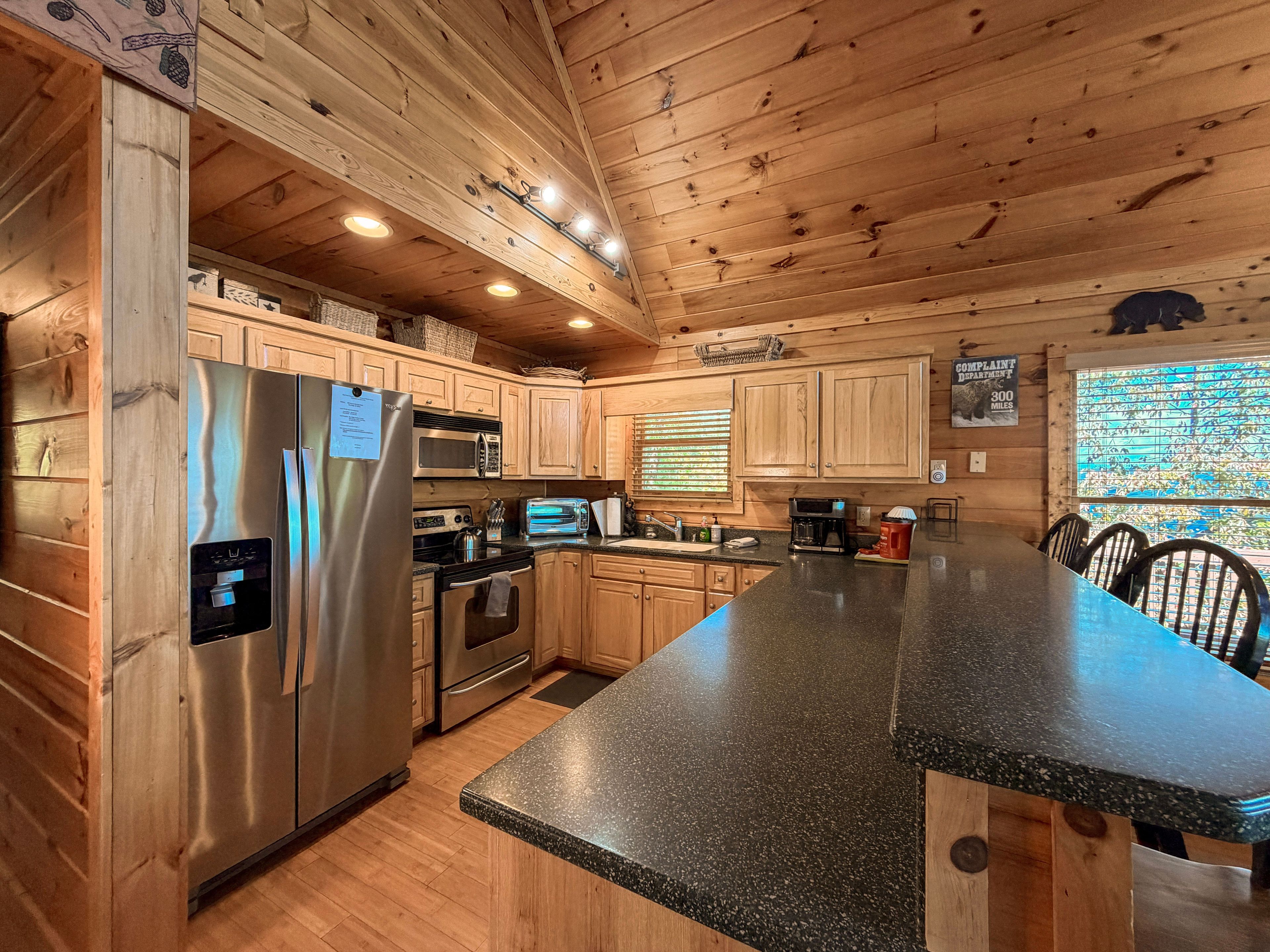 A wooden cabin kitchen with a granite countertop, stainless steel appliances, and a large window with blinds.