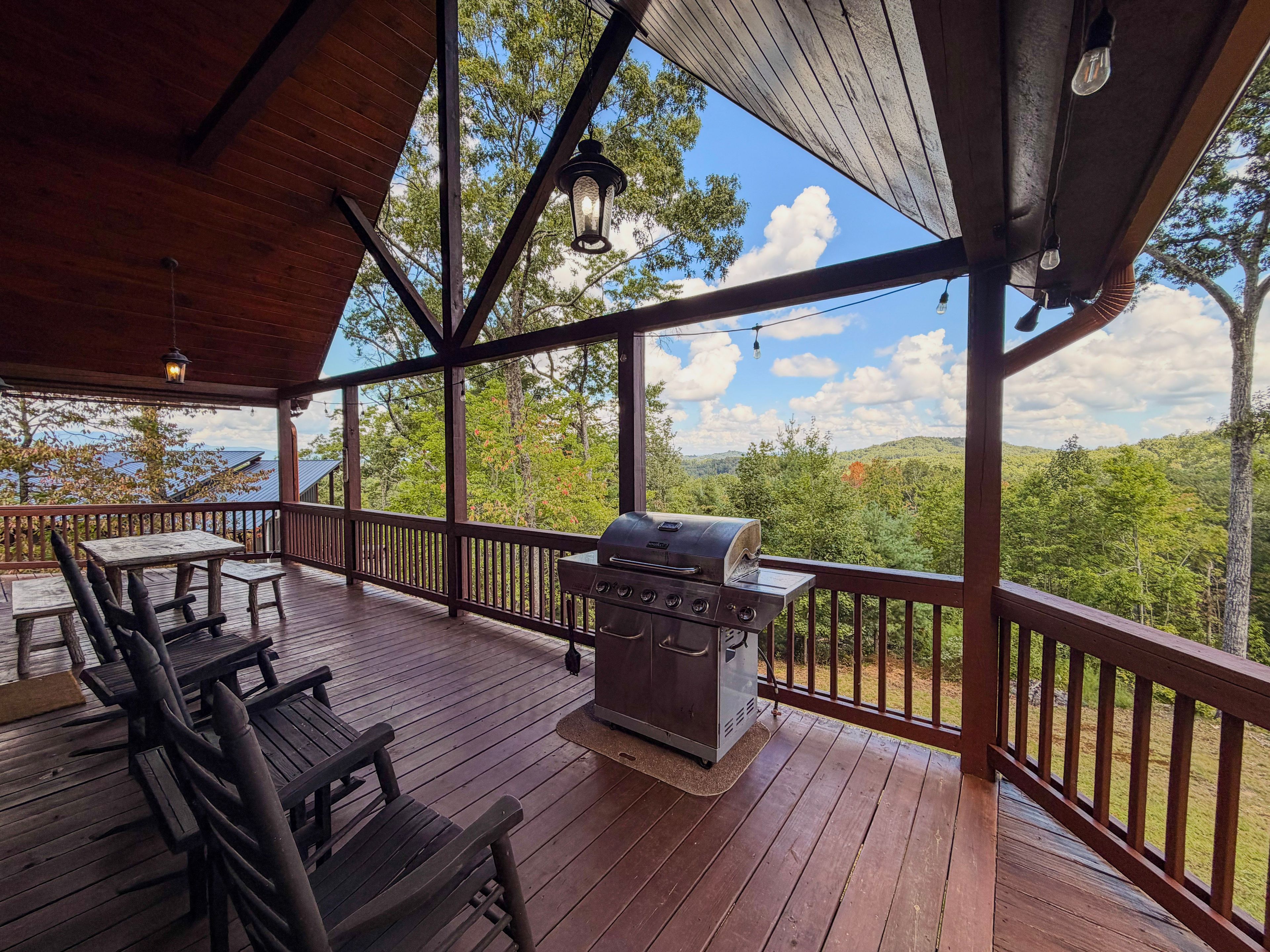 A covered outdoor deck with a grill, wooden furniture, and a view of a forested landscape.