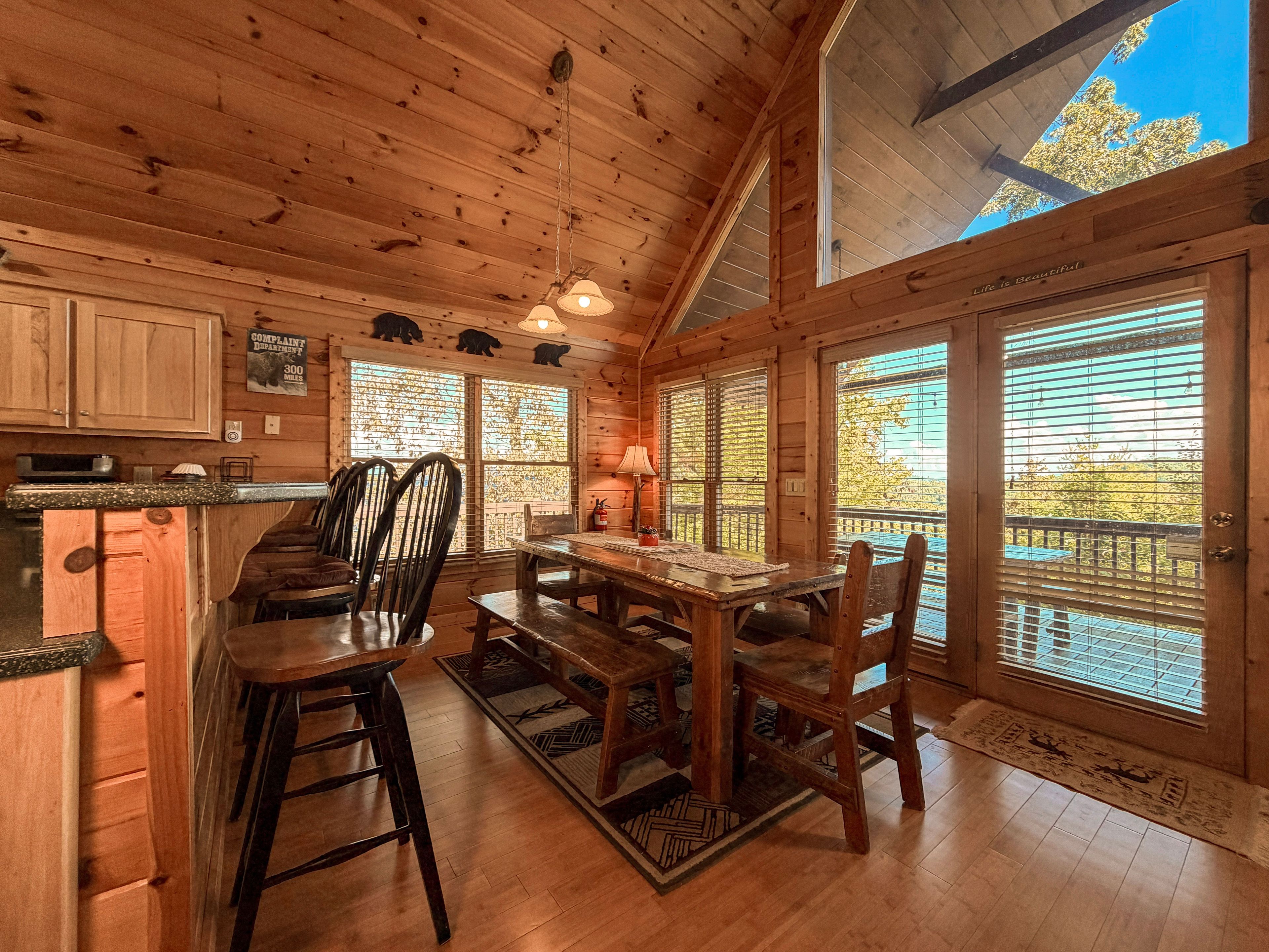 A wooden cabin interior with a dining area, kitchen, and large windows.