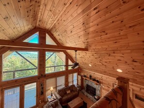 A cabin with a stone fireplace, wooden ceiling beams, and large windows offering a view of the forest. - Mountain View Cabin Near Downtown Blue Ridge (Blue Ridge)