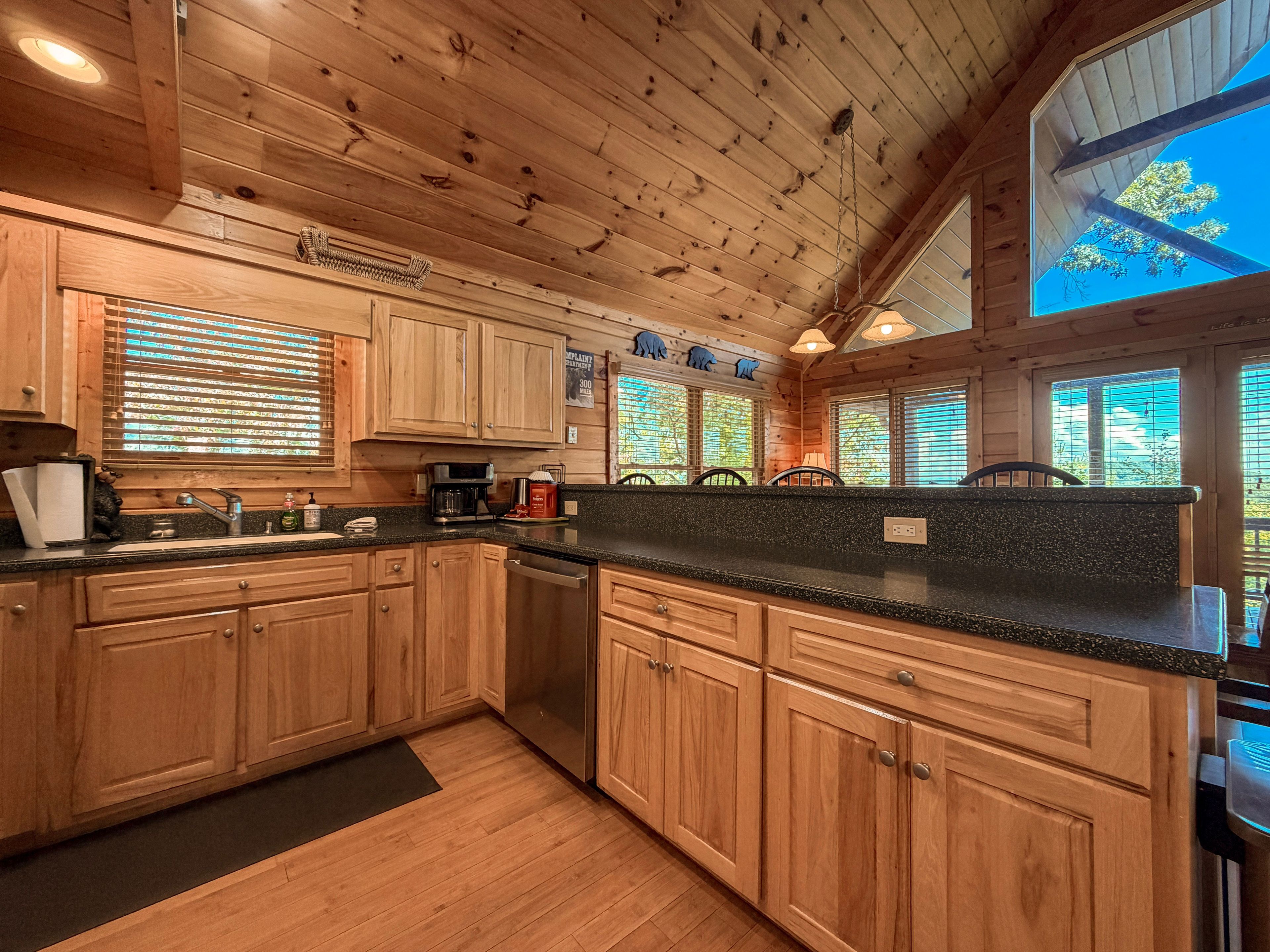 A wooden cabin kitchen with a granite countertop, wooden cabinets, and a large window with a view of trees.