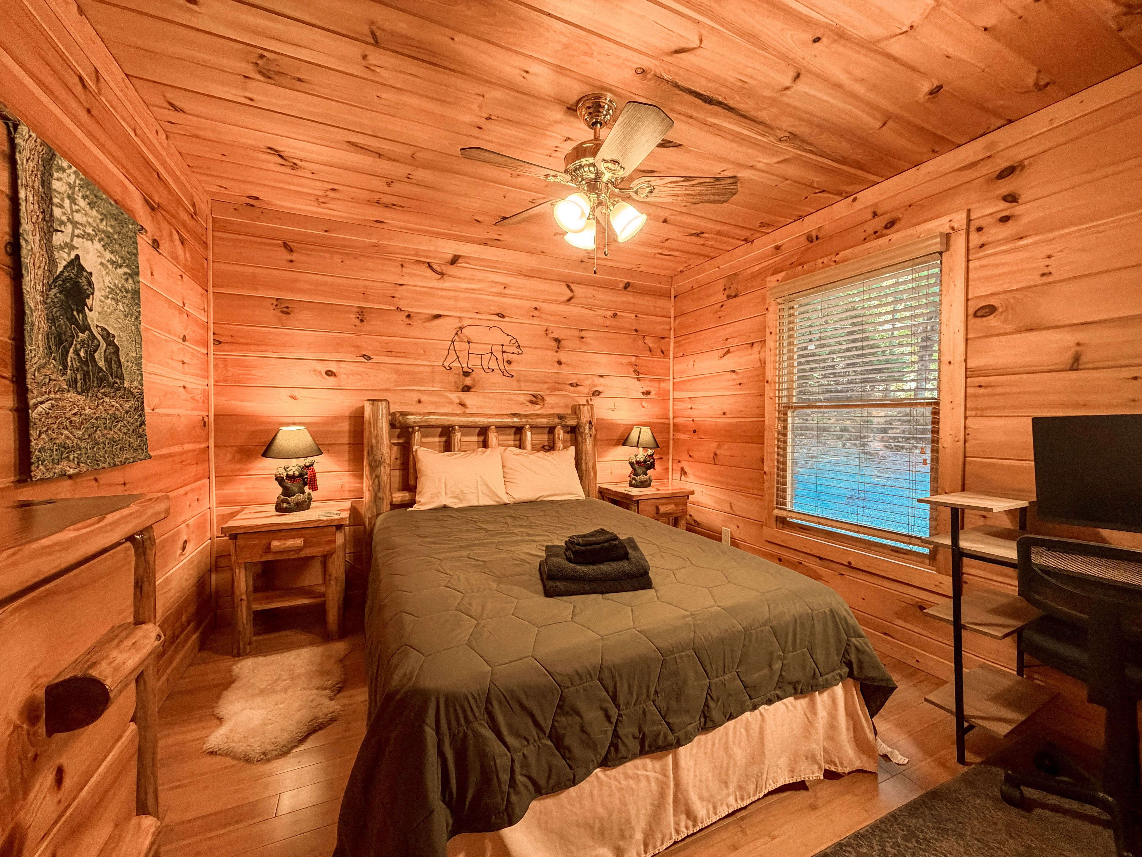 A wooden cabin bedroom with a bed, two bedside tables, a ceiling fan, a window with blinds, and a wall-mounted TV.