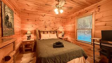 A wooden cabin bedroom with a bed, two bedside tables, a ceiling fan, a window with blinds, and a wall-mounted TV.