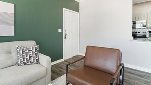 A living room with a sofa, a patterned throw pillow, a brown leather chair, and a kitchen visible through an open door. - Landing | Beautiful 1BD, Clubhouse, Gym (Katy)