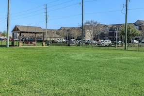A grassy area with a gazebo, a fenced parking lot, and residential buildings in the background. - Landing | Amazing 1BD, Gym, Clubhouse (Katy)