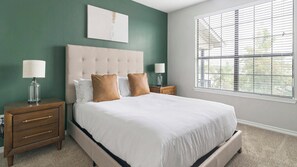 Un dormitorio con una cama grande, dos mesitas de noche, una mesita auxiliar, una ventana con persianas y una obra de arte colgada en la pared.