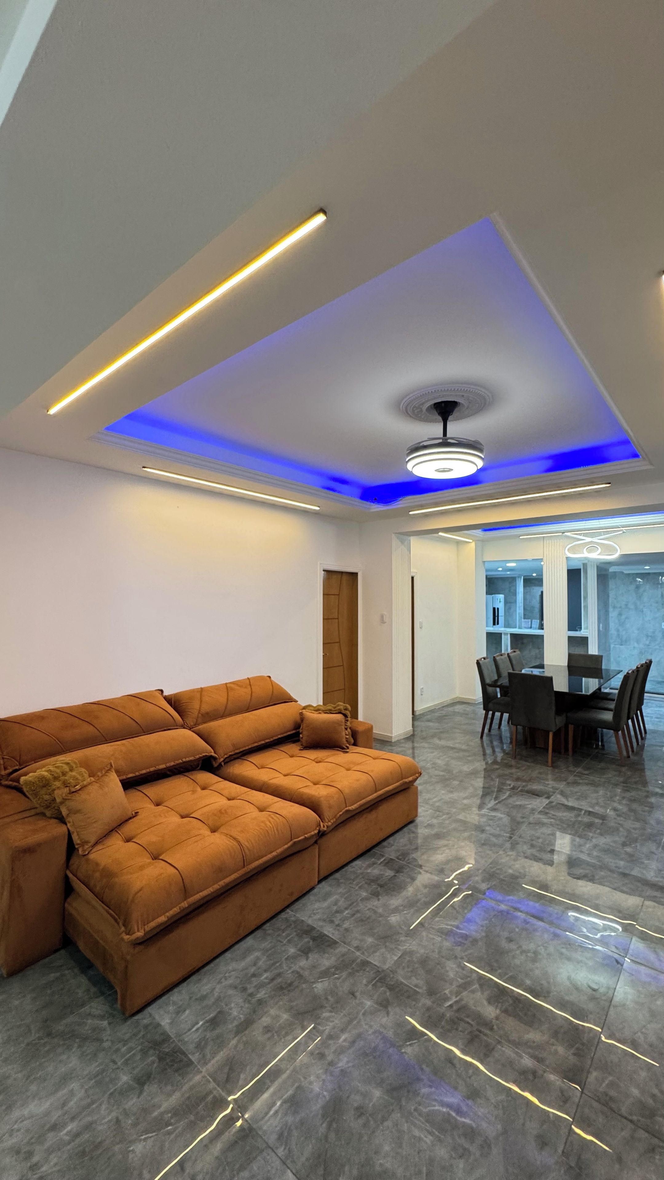 A modern living room with a sectional sofa, a dining area with a table and chairs, and a ceiling with recessed lighting and a blue accent.
