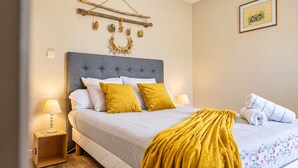 A neatly made bed with a grey headboard, white and mustard pillows, and a yellow throw blanket. There is a wooden nightstand with a lamp, a framed picture on the wall, and a small wreath above the bed. - Le Cocon du Bénitier - plage et lagon (Saint-Paul)