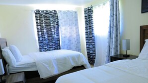 Quarto de hotel com duas camas de solteiro, mesa de cabeceira com abajur e cortinas nas janelas.