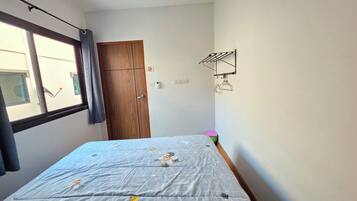 A bedroom with a bed, a wooden wardrobe, a window with curtains, a wall-mounted drying rack, and a small pink trash bin.