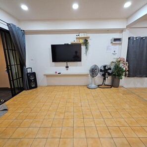 A room with a flat-screen TV mounted on the wall, a small shelf with decorative items, a potted plant, and a speaker.
