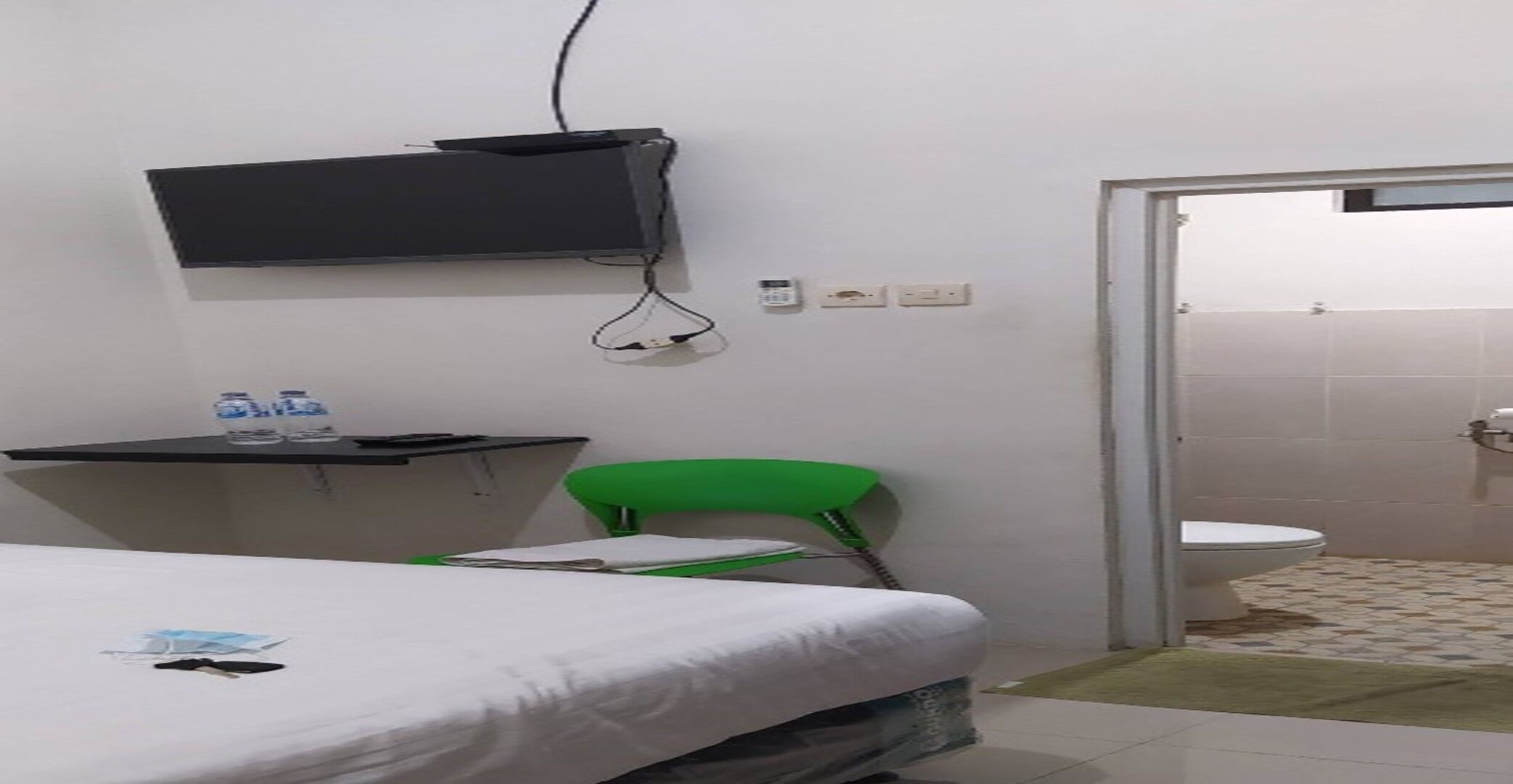 Una habitación de hotel pequeña con una cama, una silla verde, un escritorio con televisor de pantalla plana y un baño visible a través de una puerta abierta.