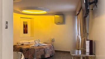 A hotel room with a bed, a desk, a chair, a flat-screen TV mounted on the wall, and a ceiling light fixture.