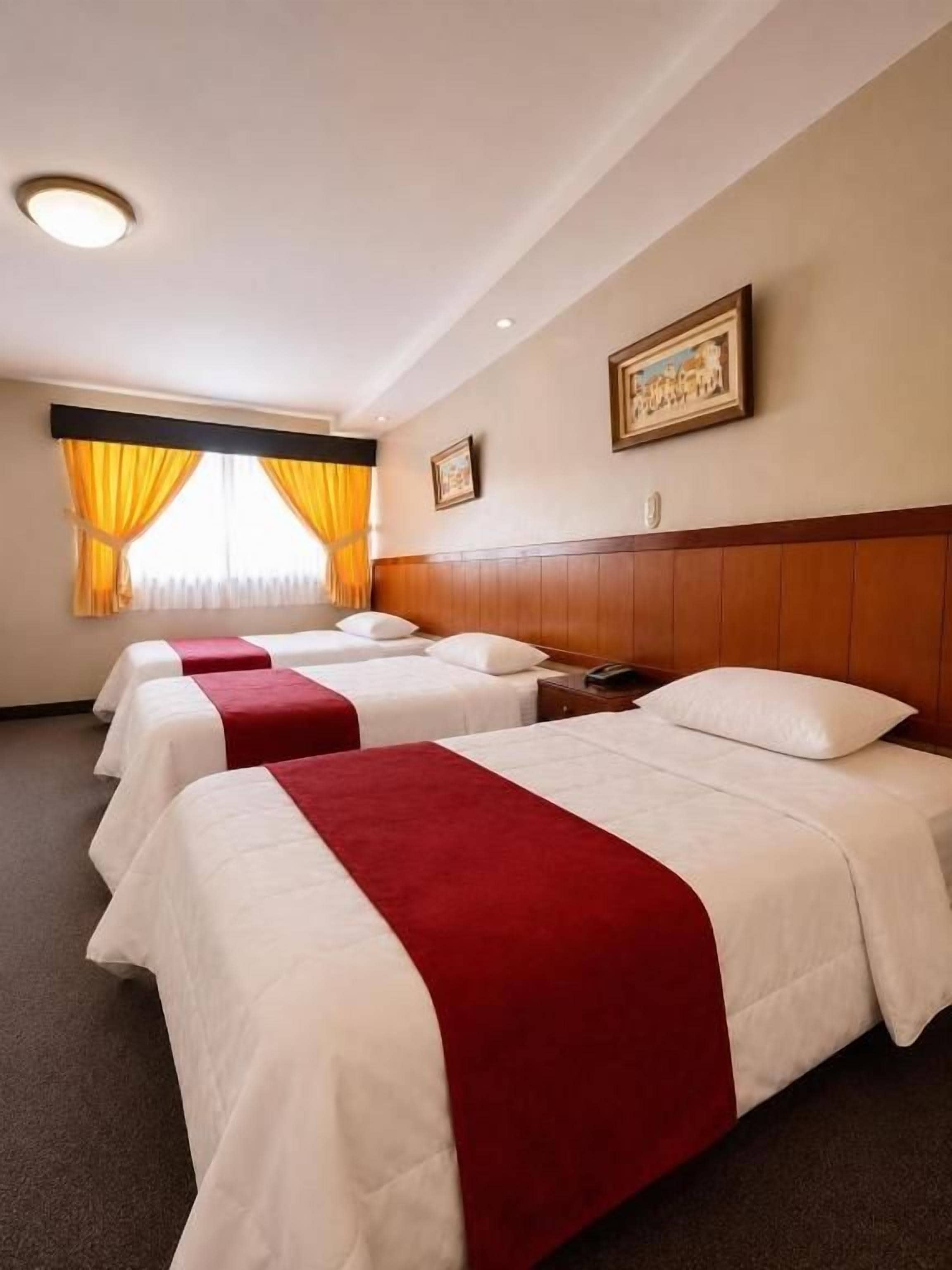 A hotel room with three beds, each with a red and white bedspread, a wooden headboard, and a painting on the wall.
