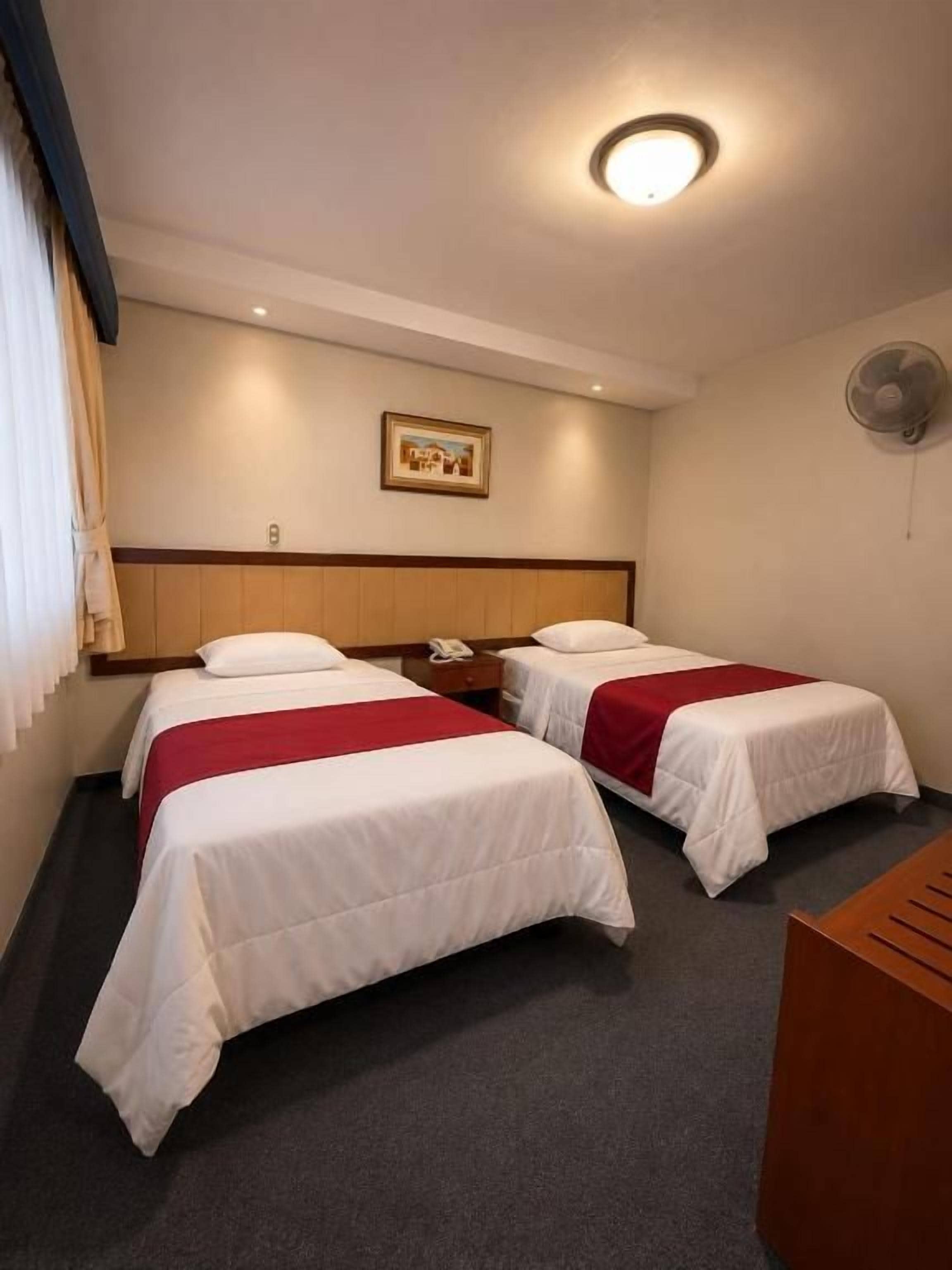 Une chambre d’hôtel avec deux lits, une tête de lit en bois, une table de chevet avec un téléphone, un ventilateur fixé au mur et un tableau encadré.