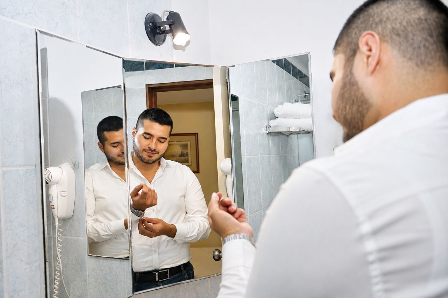 A man in a white shirt using a hair dryer in a bathroom with a mirror, a towel rack, and a wall-mounted light fixture.