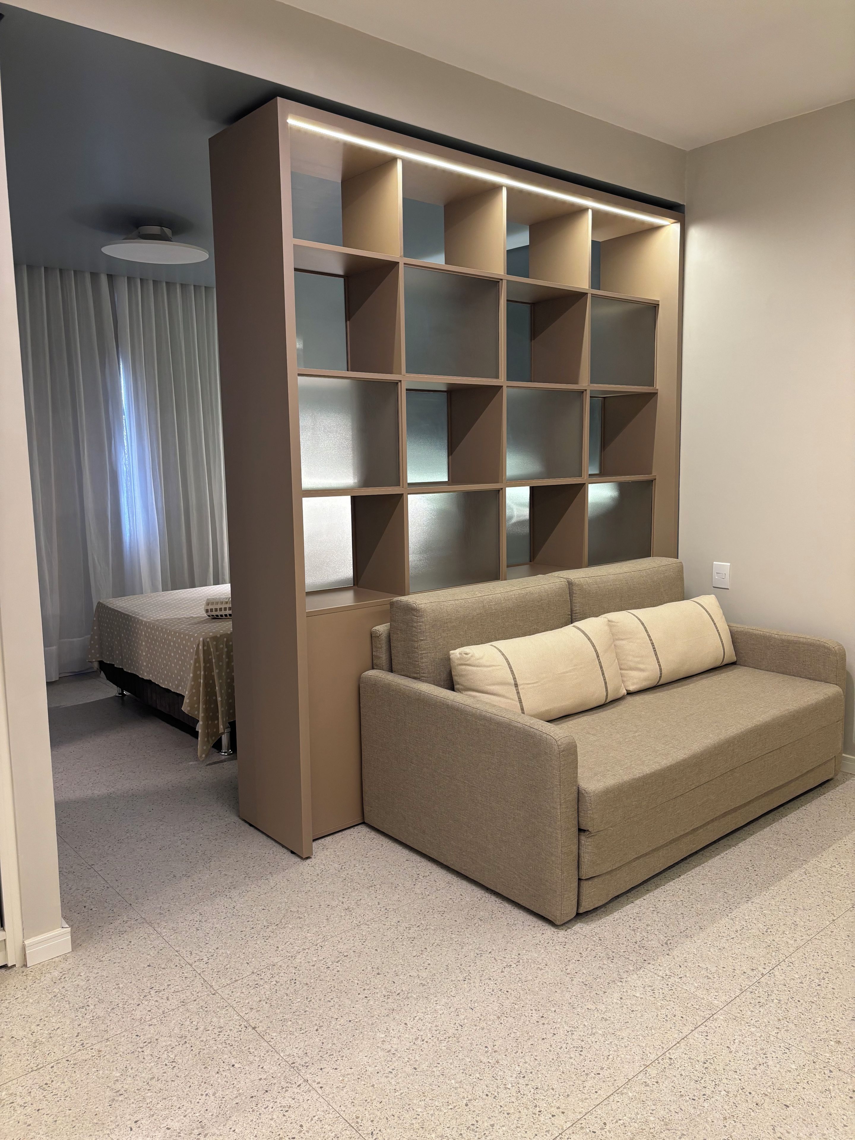 A modern living room with a beige sofa, a built-in shelving unit, and a bed with a patterned bedspread.