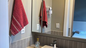 Ein Badezimmer mit einem weißen Waschbecken, einem Handtuchhalter mit roten Handtüchern, einem Föhn und einem Spiegel, der ein Bett reflektiert.