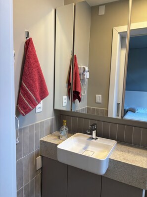 Un baño con un lavamanos blanco, un perchero con toallas rojas, un secador de pelo y un espejo que refleja una cama.