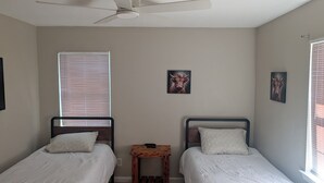 Une chambre avec deux lits simples, un ventilateur de plafond et deux tableaux accrochés au mur.