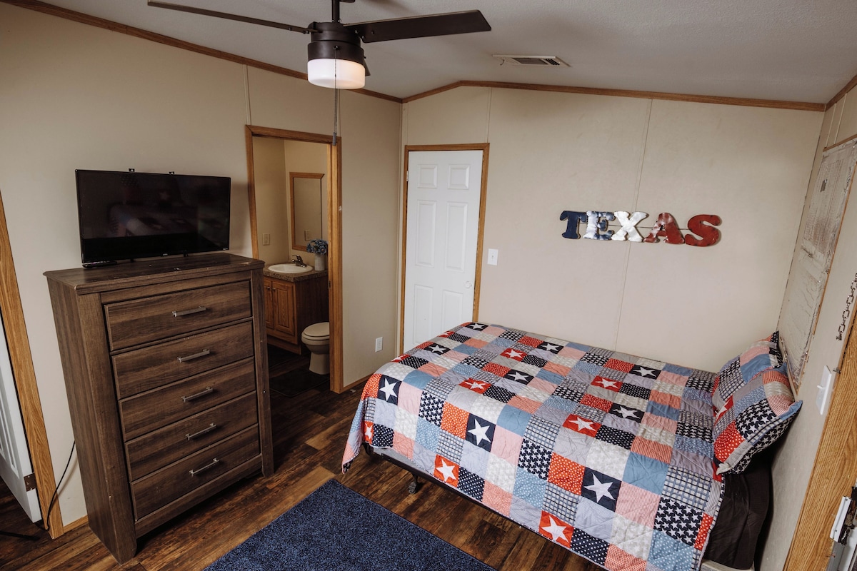 A bedroom with a wooden dresser, a television, a bed with a quilt, and a door labeled 'TEXAS'.