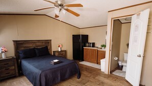 A bedroom with a bed, nightstands, a ceiling fan, a refrigerator, a microwave, a small cabinet, and a bathroom door. - 302 Lodging (Kermit)