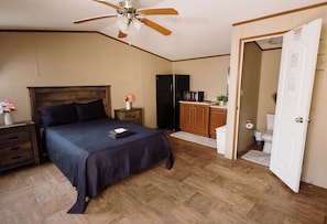 A bedroom with a bed, nightstands, a ceiling fan, a refrigerator, a microwave, a small cabinet, and a bathroom door. - 302 Lodging (Kermit)