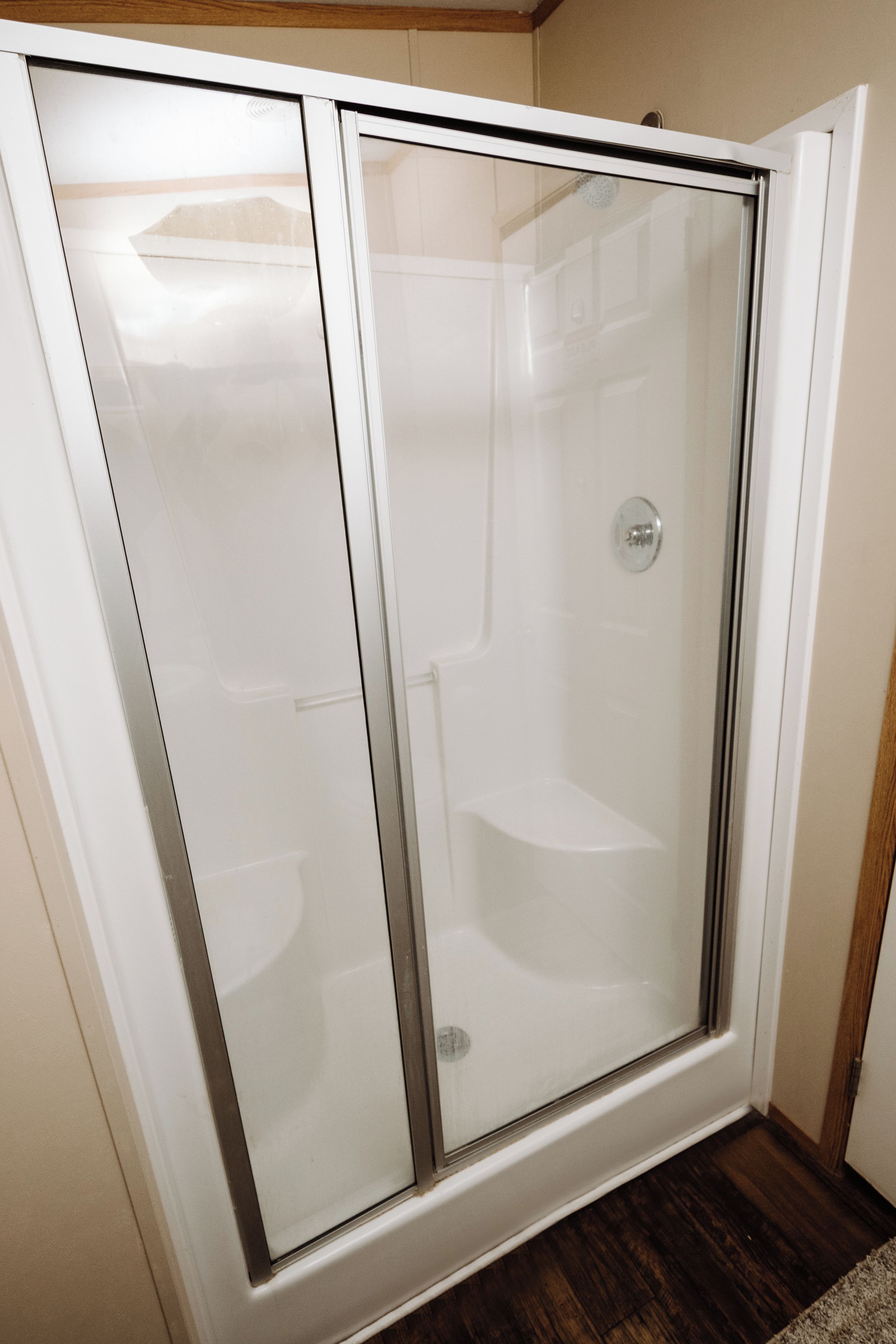 A glass-enclosed shower stall with a white frame, a visible drain, and a wooden floor.