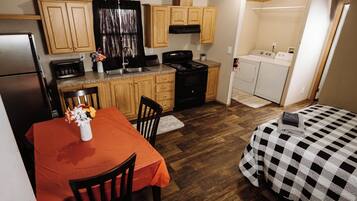 A compact kitchen with wooden cabinets, a dining table with a red tablecloth, and a bed with a checkered bedspread.