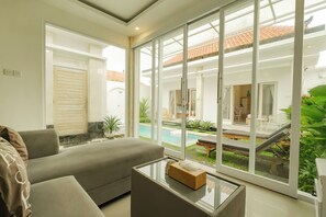 A modern living room with a sofa, a glass coffee table, and a view of a swimming pool and outdoor seating area. - Villa Warna Canggu (Badung)