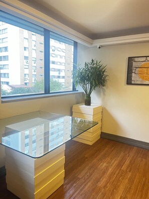 A room with a glass-top table, a potted plant, a framed picture on the wall, and a view of a building through a large window.