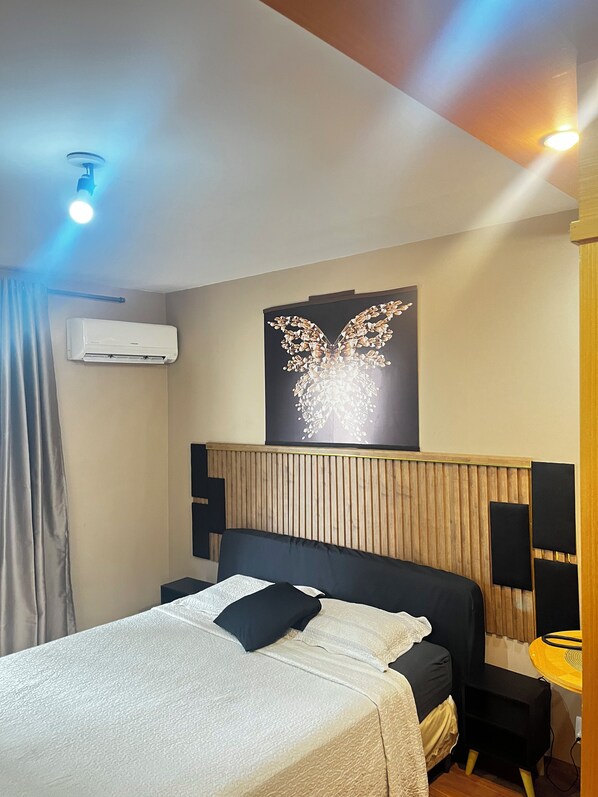 A hotel room with a bed, a wall-mounted air conditioner, a decorative wall piece, and a bedside table with a lamp.