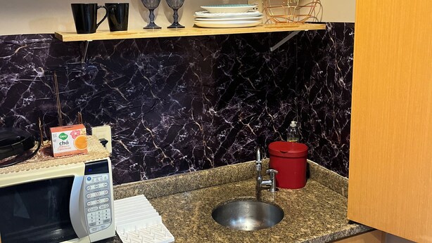 A compact kitchen with a microwave, sink, and a shelf holding cups and glasses. - Cozy 1-bedroom bed & breakfast in peaceful São Paulo with AC (São Paulo)