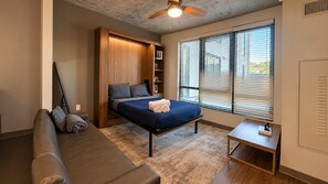Ein Schlafzimmer mit einem Bett, einem Einbauschrank, einem Fenster mit Jalousien, einem kleinen Tisch und einem Deckenventilator.