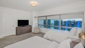 A modern hotel room with a large window, a flat-screen TV, a wooden dresser, a beige armchair, and a white bed with a gold lamp. - Luxury Four seasons Hotel Miami 2 Bedroom Oceanview Luxury Suite. (Miami)