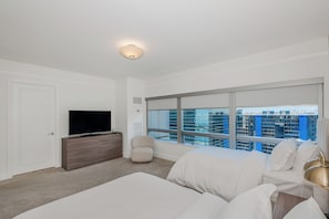 Quarto de hotel moderno com uma janela grande, televisão de tela plana, cômoda de madeira, poltrona bege e cama branca com abajur dourado.