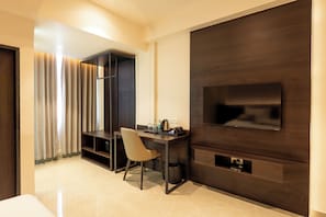 A hotel room with a dark wood panel wall, a flat-screen TV mounted on it, a desk with a chair, and a small trash bin.