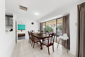A dining area with a large table and chairs, a clock on the wall, and a view through a sliding glass door. - Spacious 4-Bed House Near Mandurah (Dudley Park)