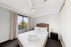 A hotel room with a bed, a nightstand, a lamp, a ceiling fan, and a window with curtains. - Spacious 4-Bed House Near Mandurah (Dudley Park)