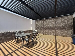 Eine Terrasse im Freien mit Tisch und Stühlen, einer gefliesten Wand und einer Metall-Dachkonstruktion.
