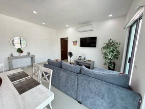 Ein modernes Wohnzimmer mit einem grauen Sofa, einem Esstisch mit Stühlen, einem flachbildfernseher an der Wand und einer Zimmerpflanze.